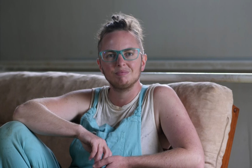 A person with blonde hair and glasses in blue overalls sitting on a couch.