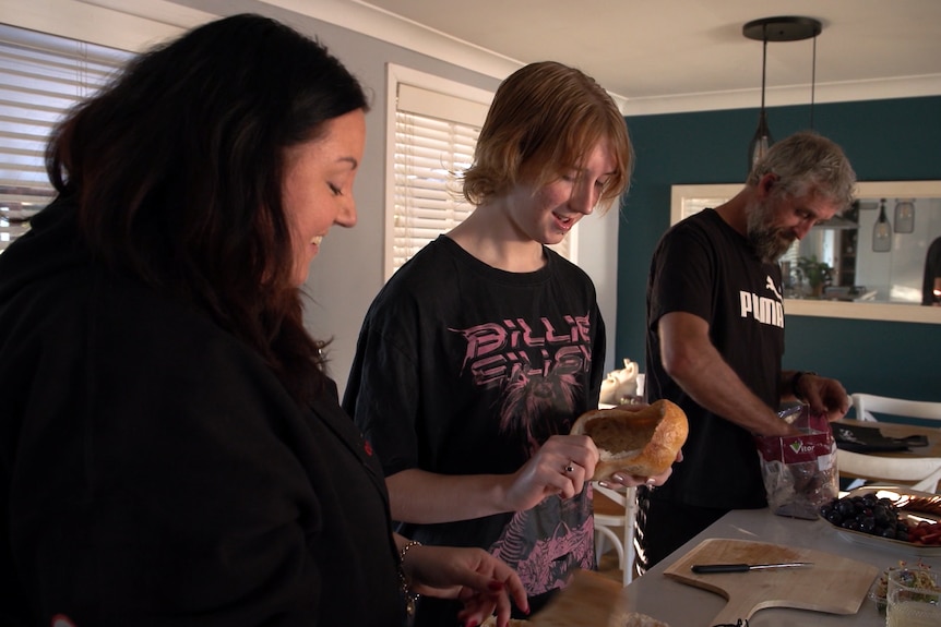 A teenage girl wearing a Billie Eilish t-shirt and her parents stand at a kitchen counter making a meal.