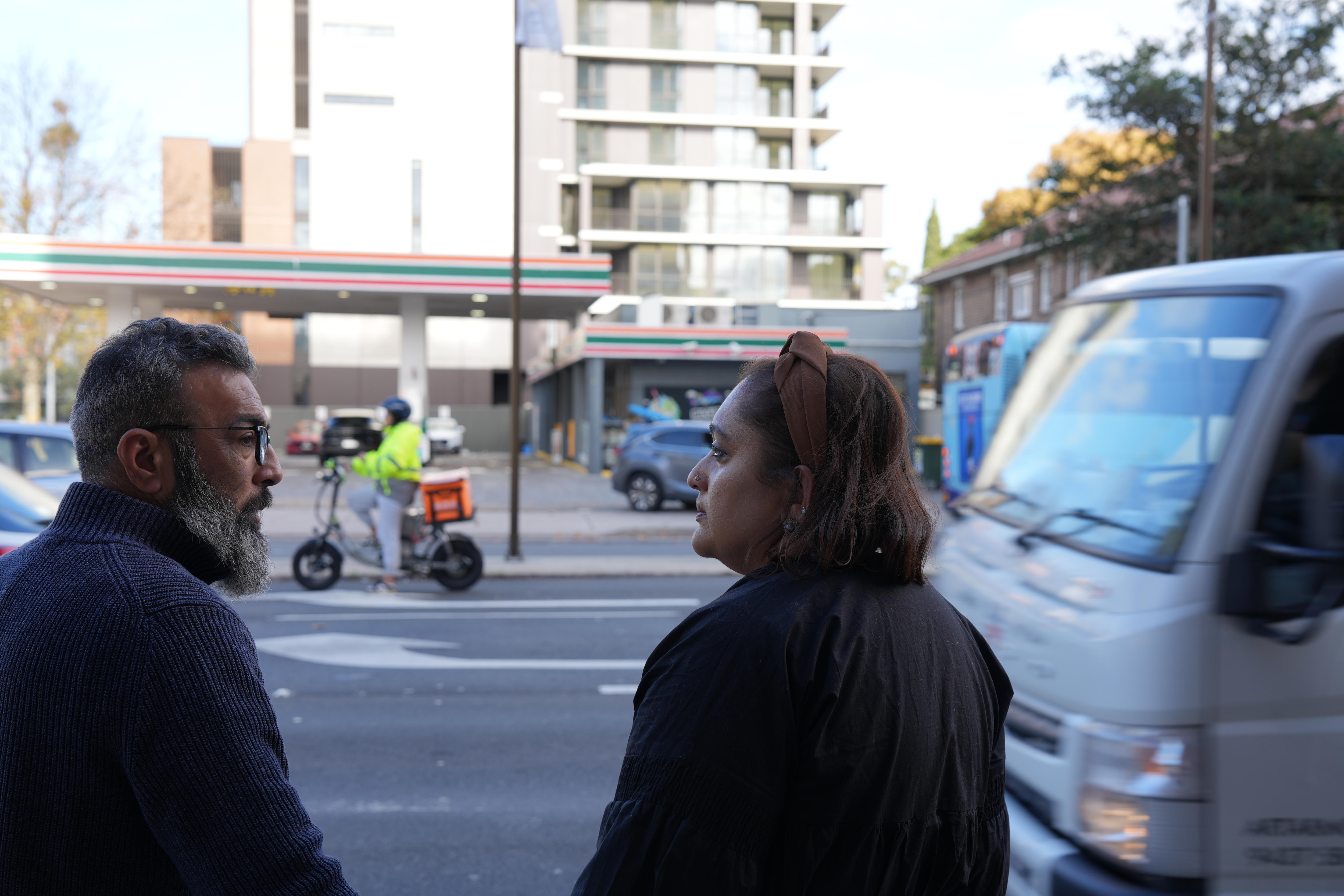 A bearded man and dark-haired woman stand across the street from a 7-Eleven petrol station.