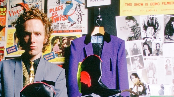 Picture of Malcolm McLaren holding a cowboy boot with posters from the 1950s behind him