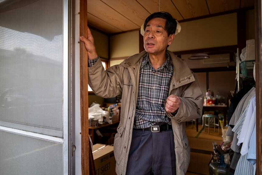 Mr Masatosi Mitani is standing in a doorway. He is wearing a beige coat and a checkered shirt.