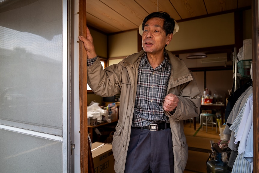Mr Masatosi Mitani is standing in a doorway. He is wearing a beige coat and a checkered shirt.