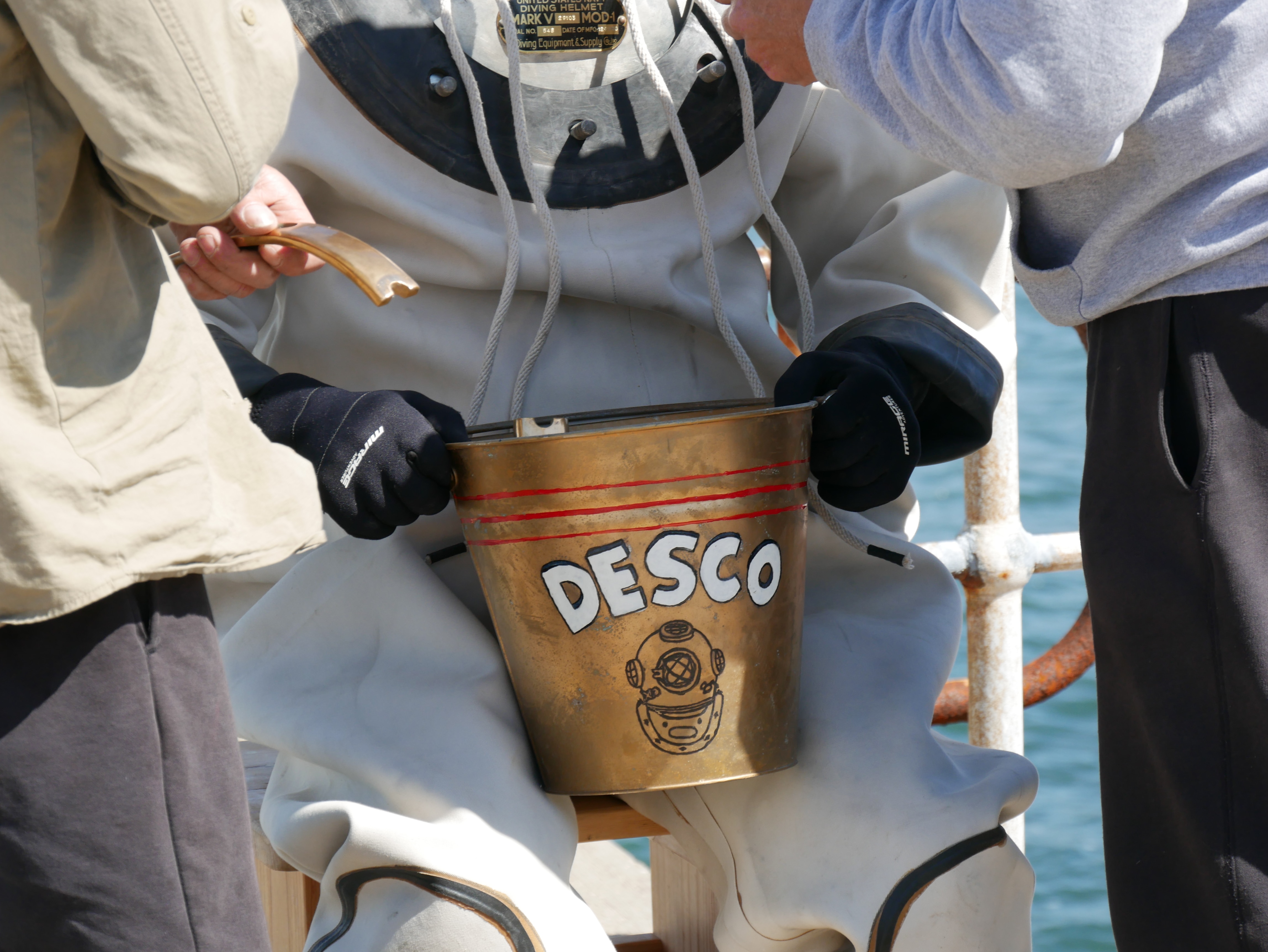 Close-up of two hands wearing black gloves holding a copper bucket with the word DESCO and a picture of a helmet. 