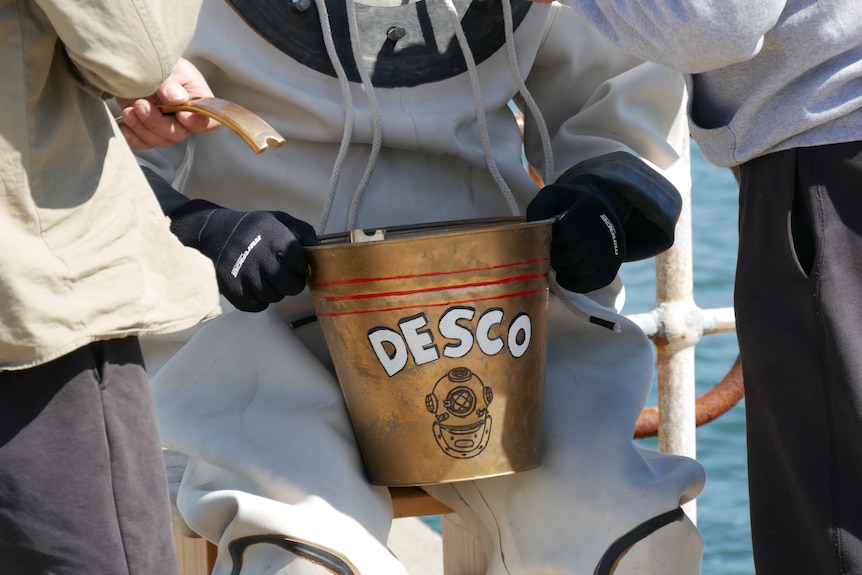 Close-up of two hands wearing black gloves holding a copper bucket with the word DESCO and a picture of a helmet. 
