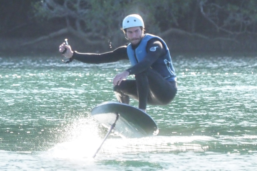 A man in a wetsuit, wearing a helmet, riding a foil board, lifted above the surface of the water on the keel.