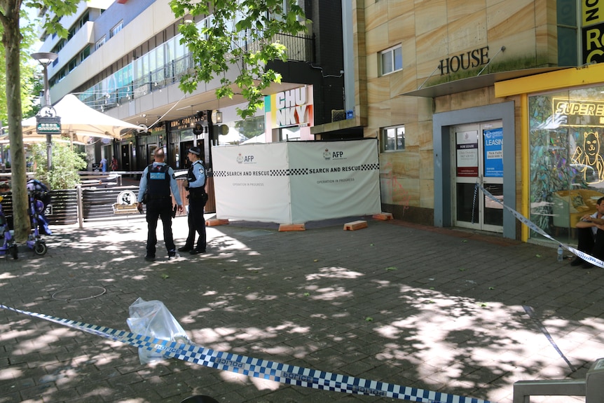 two police stand next to police tape and white shields in a laneway with shops