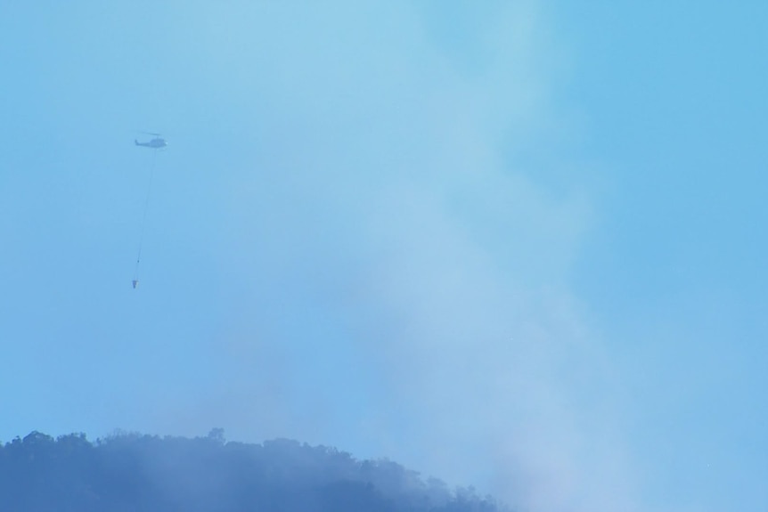 A chopper carrying water flies over a mountain with bushfire smoke