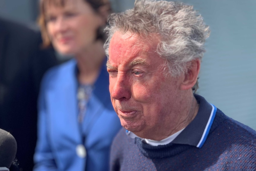 A man in a blue jumper with grey hair addresses a microphone.