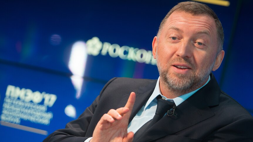 Oleg Deripaska wearing a suit in front of a blue background mid-sentence while gesturing with his right hand