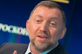 Oleg Deripaska wearing a suit in front of a blue background mid-sentence while gesturing with his right hand