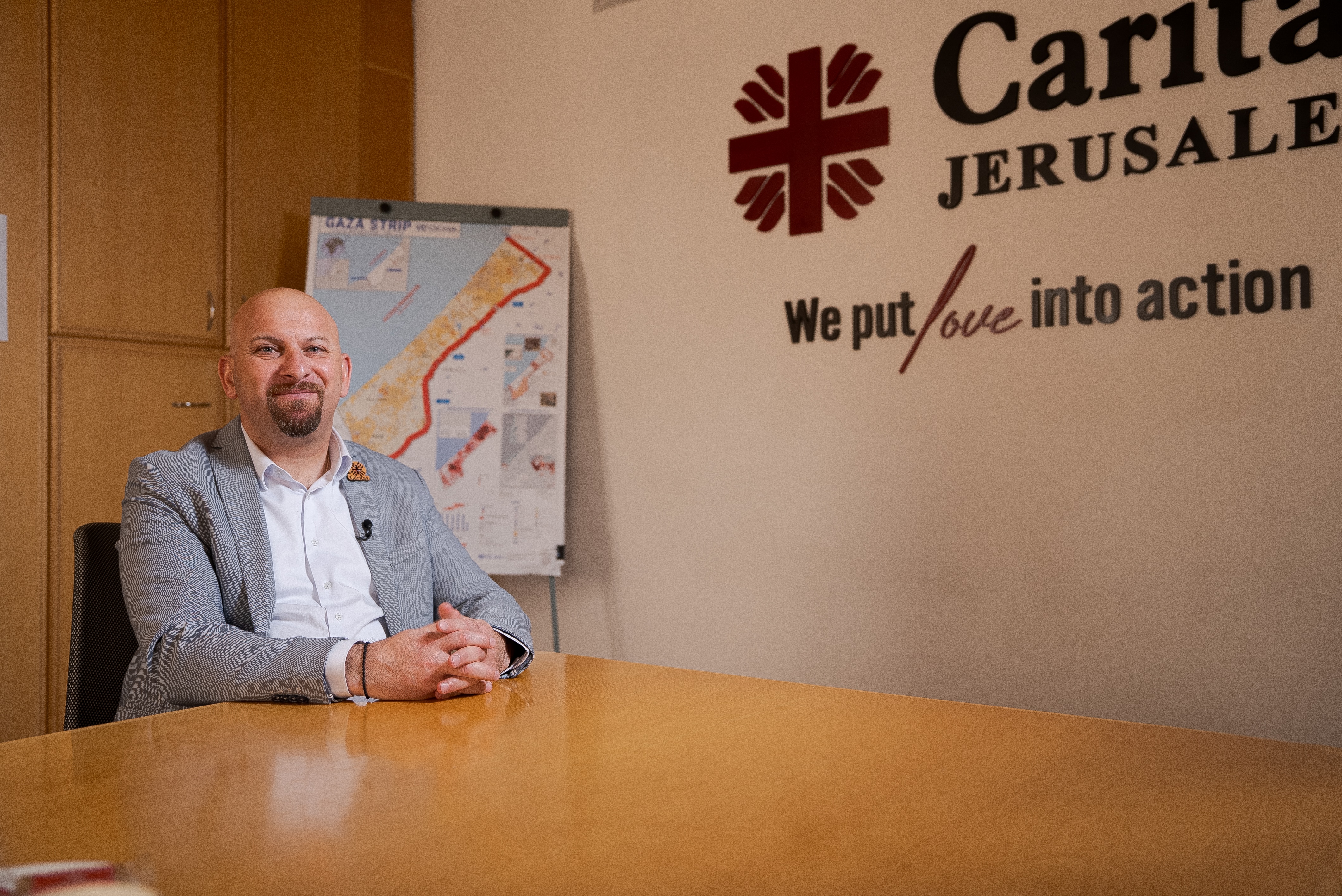 Anton Asfar wearing a suit sitting at a desk with a map of Gaza on a poster behind him and Caritas Jerusalem logo on the wall