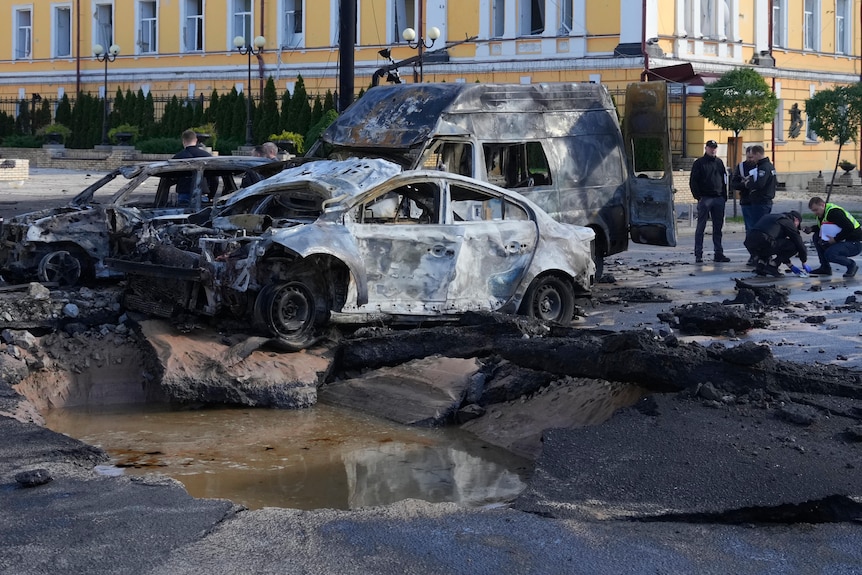 Damaged cars and a chunk of road is blown up in a city street