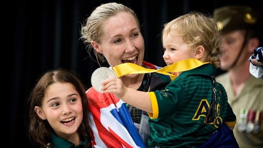A woman with blonde hair holds her daughter, who is holding a medal. Her other daughter is also in the picture.