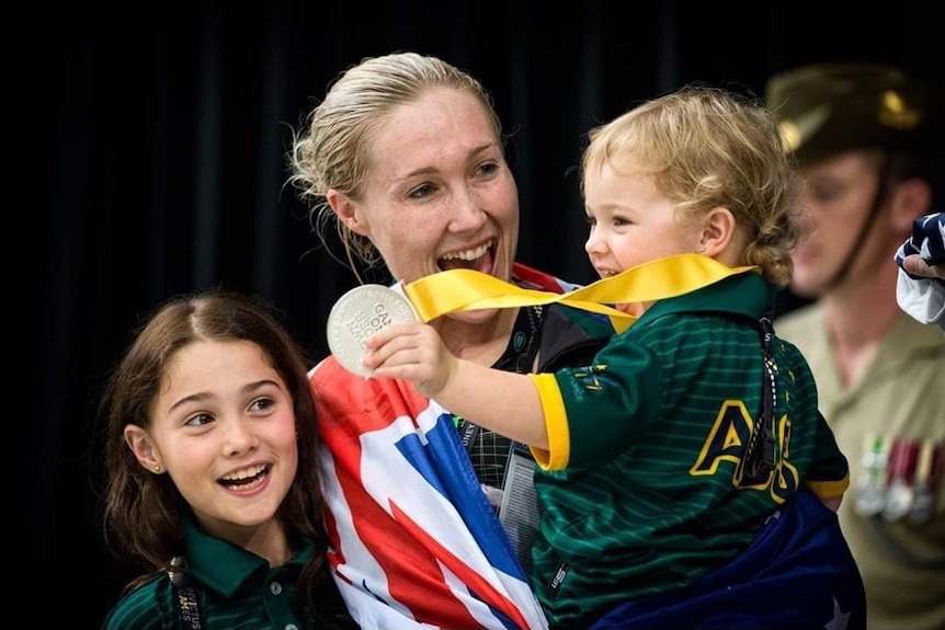 A woman with blonde hair holds her daughter, who is holding a medal. Her other daughter is also in the picture.