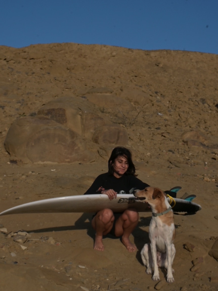 A woman crouches on the beach with a surfboard on her lap, she pats a dog
