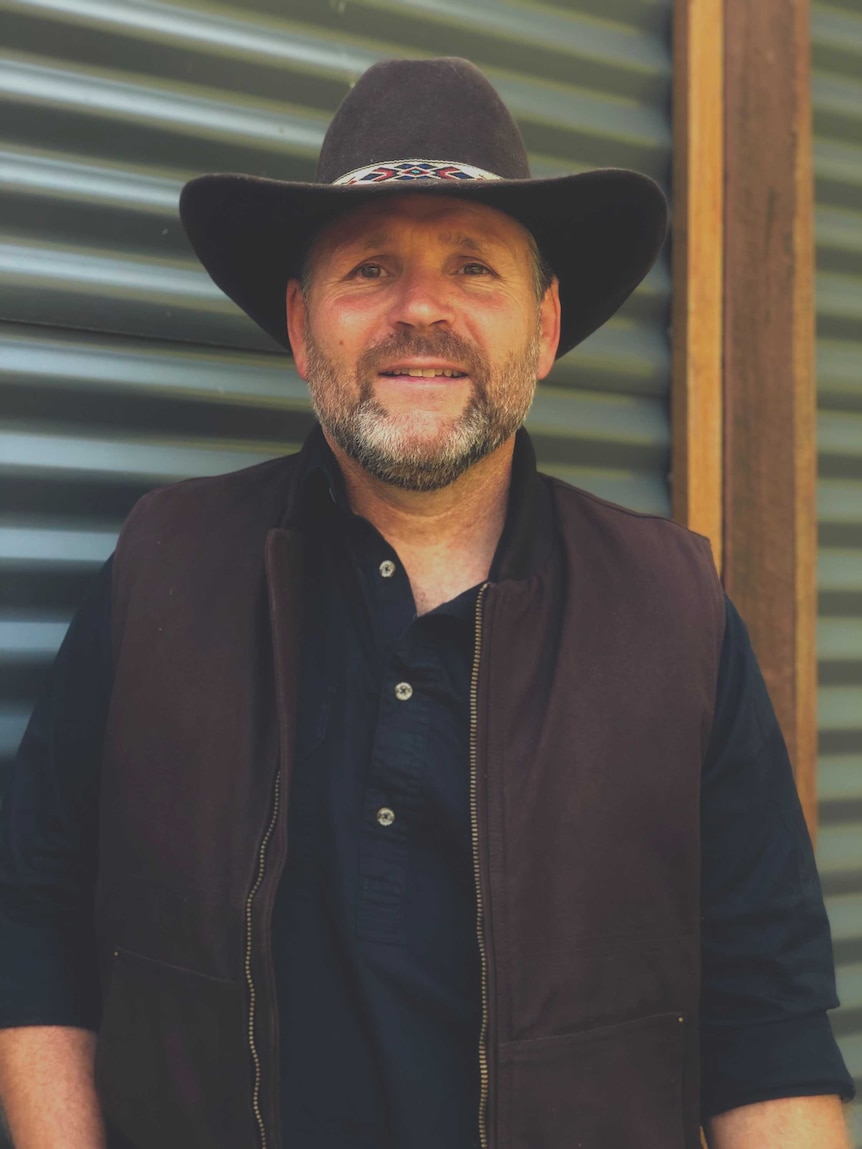 A man in a cowboy-style hat, shirt and vest smiles to camera.