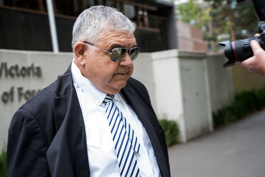 Man in aviator sunglasses and  suit, on right of frame camera and hand can be seen.