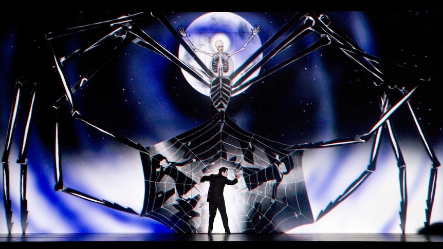 A woman has the image of a giant spider projected over her as she sings. A man below her is caught in a web.