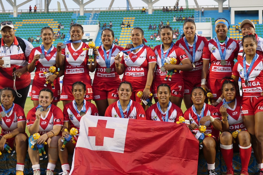 A group of women wearing Tonga jerseys hold up their flag and smile for the camera.