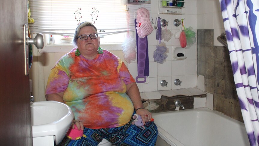 A woman sitting in a worn-down bathroom. Some of the tiles have fallen off the walls.