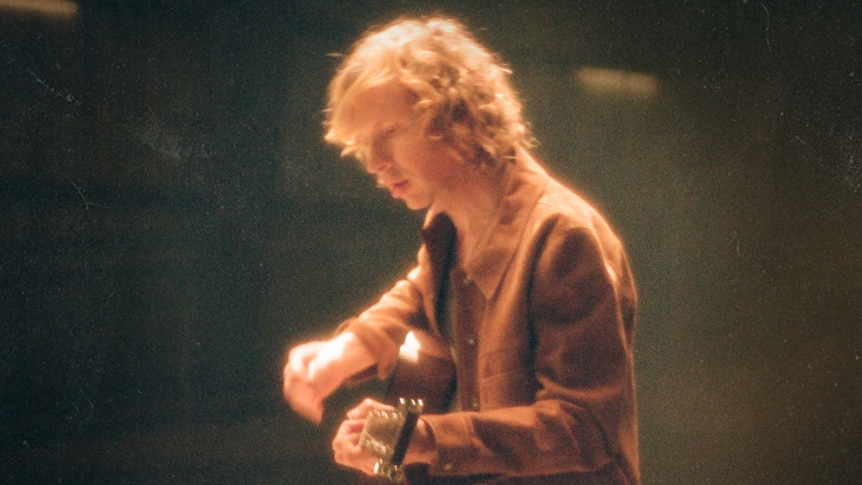Beck sits on a stool and plays an acoustic guitar. He wears a brown jacket.