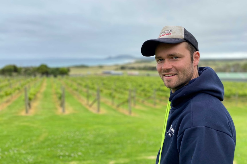 A man wearing a hoodie, cap and sporting some stubble stands in a green field.