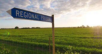 A sign points the way towards Greenough Regional Prison in front of a lush green field and sunrise.