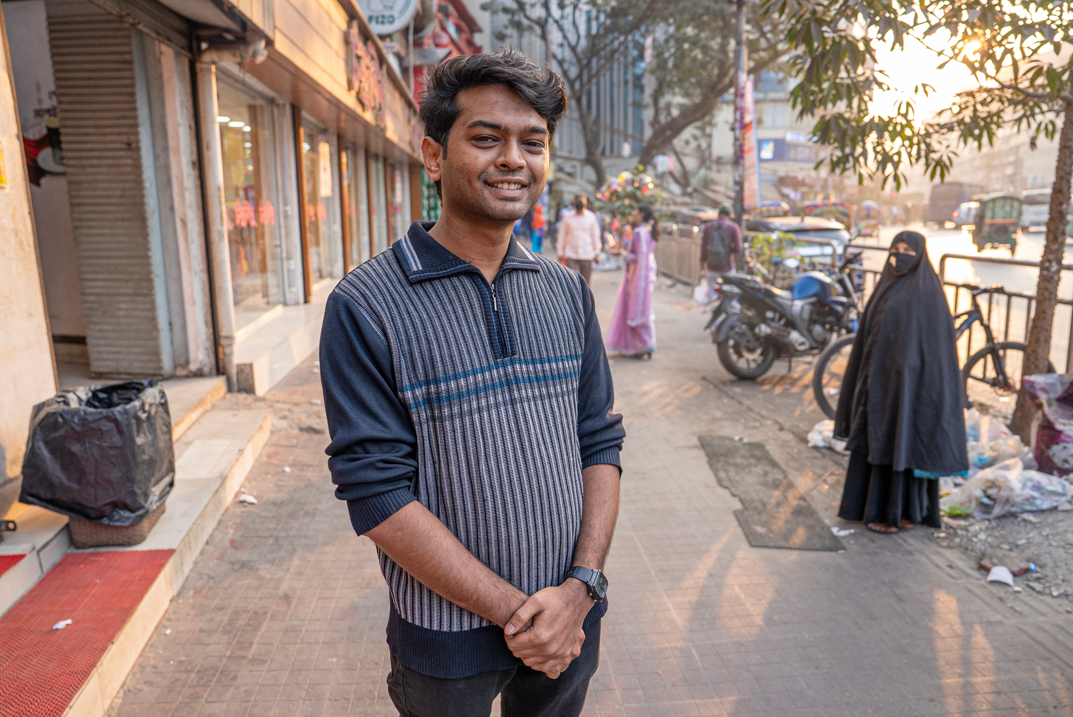 A Bangladesh man smiles on a sidewalk with a striped shirt on.