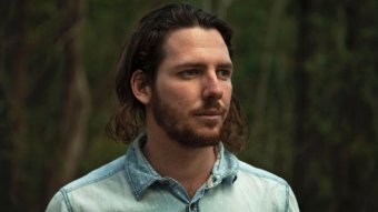 A man with long brown hair wears a denim shirt.