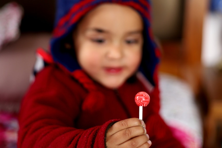 A young boy in a red jacket holds a red swirl lollipop in front of him.