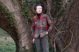Princess Anne in a check fleece shirt stands next to a tree 