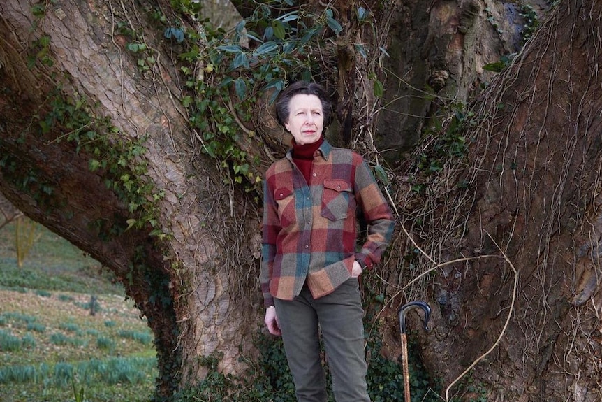 Princess Anne in a check fleece shirt stands next to a tree 