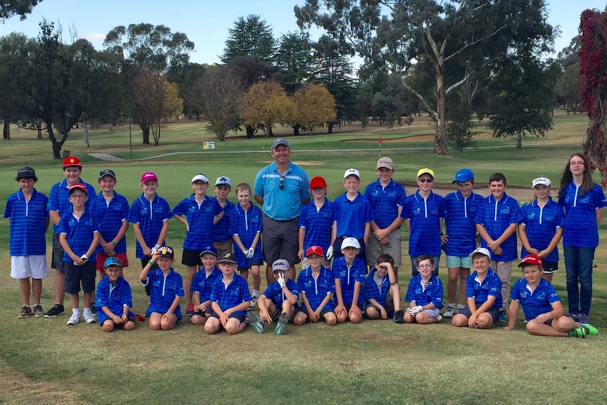 AA smiling man with a bunch of about 20 younger people stand on a golf course. All are wearing blue shirts