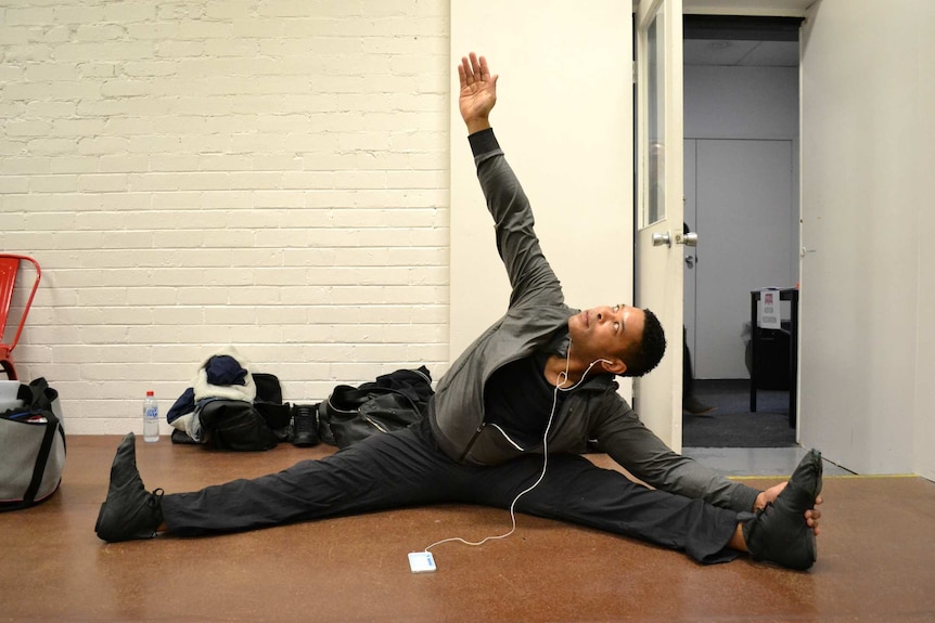 A man stretches while listening to music on his phone in a dance studio.