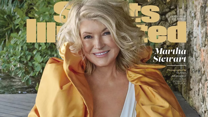 Martha Stewart, wearing short blonde hair, a white swimsuit and gold wrap, poses for a photo, smiling