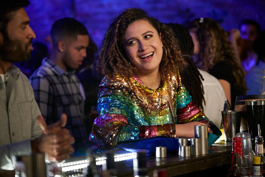 Rose Matafeo grins while leaning across a bar in a nightclub wearing a sequinned dress in Starstruck