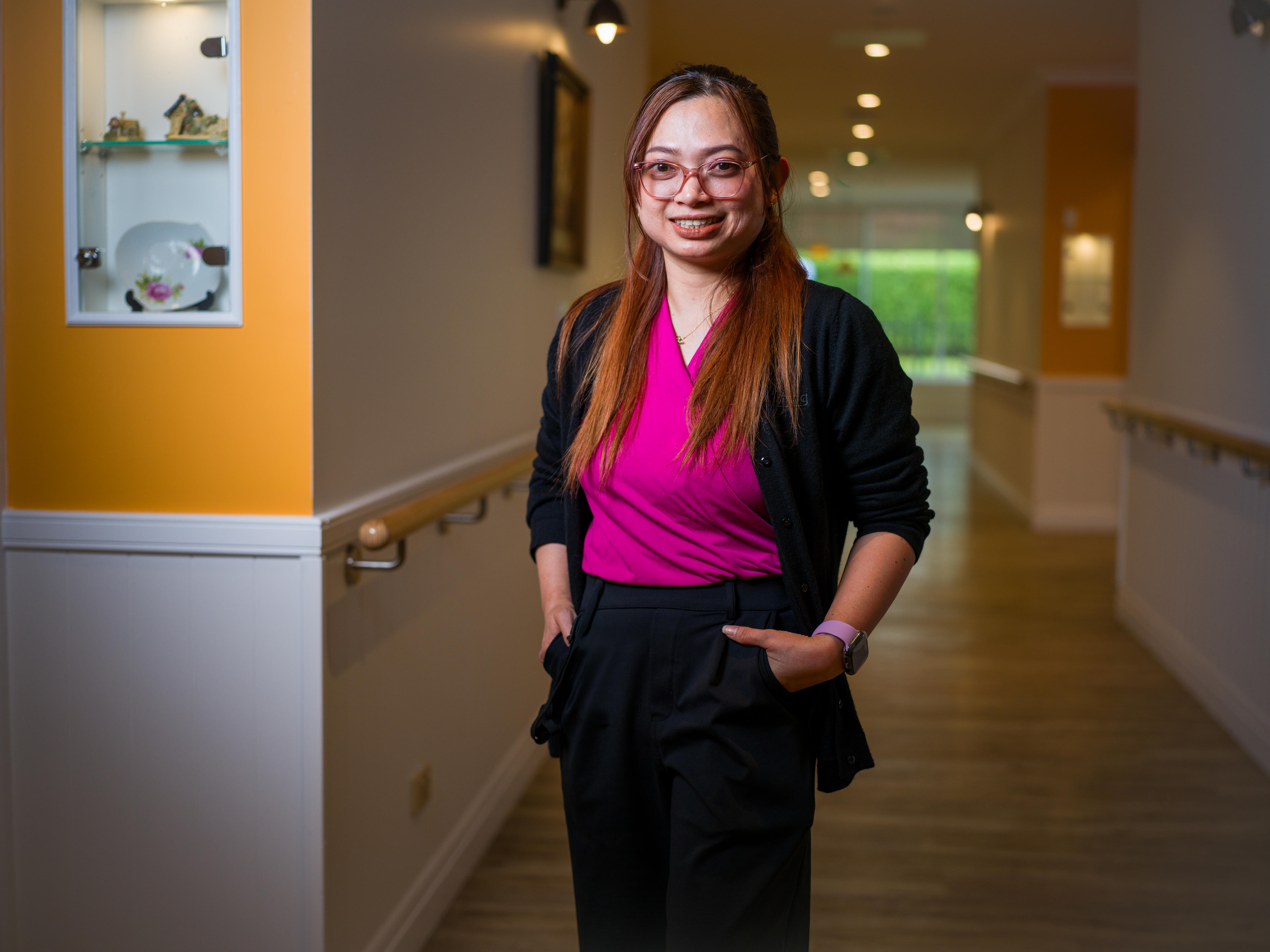 A woman with long brown hair and pink glasses stands in a residential aged care facility hallway.