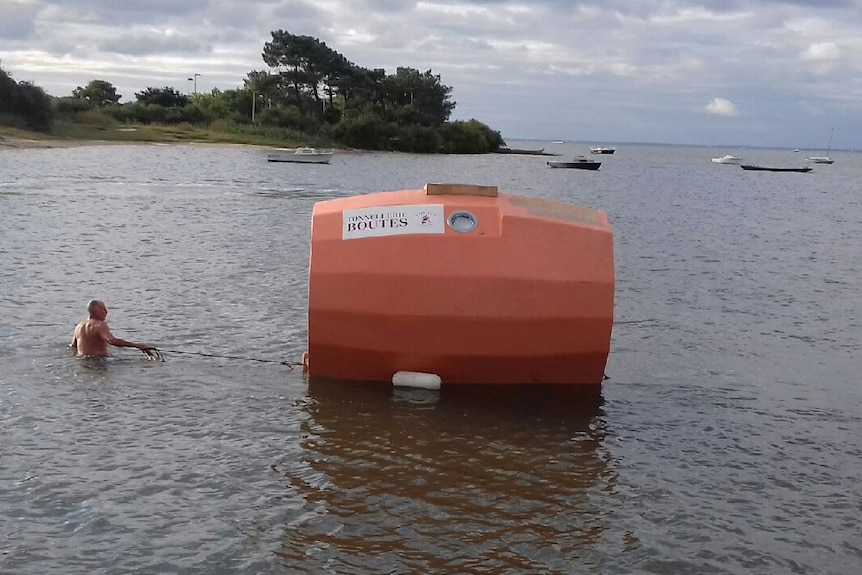 A shirtless Jean-Jacques Savin pulls orange barrel towards the shore with a rope on an overcast day.