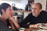 A couple in their 40s sit at their kitchen table, smiling at each other.