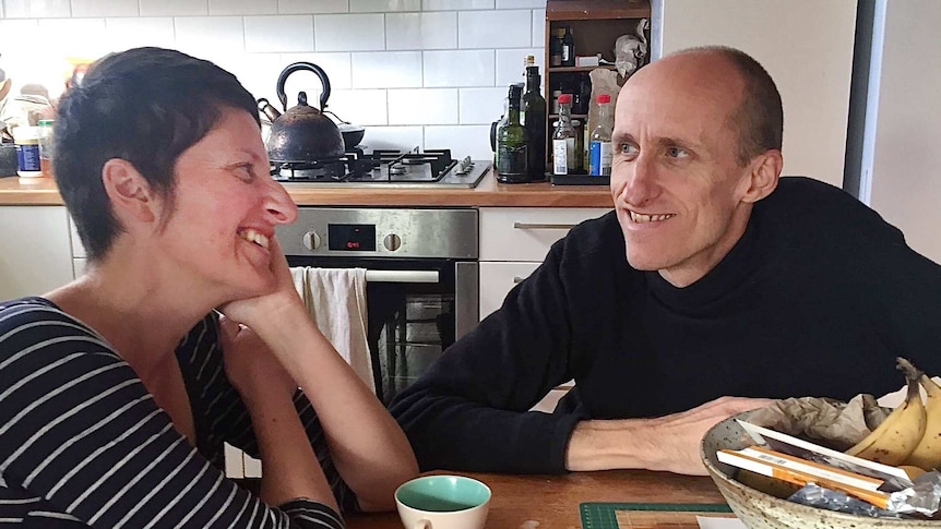 A couple in their 40s sit at their kitchen table, smiling at each other.