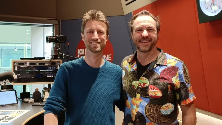 a man in a blue shirt poses with a man in a floral hawaiian shirt for the camera in a radio studio