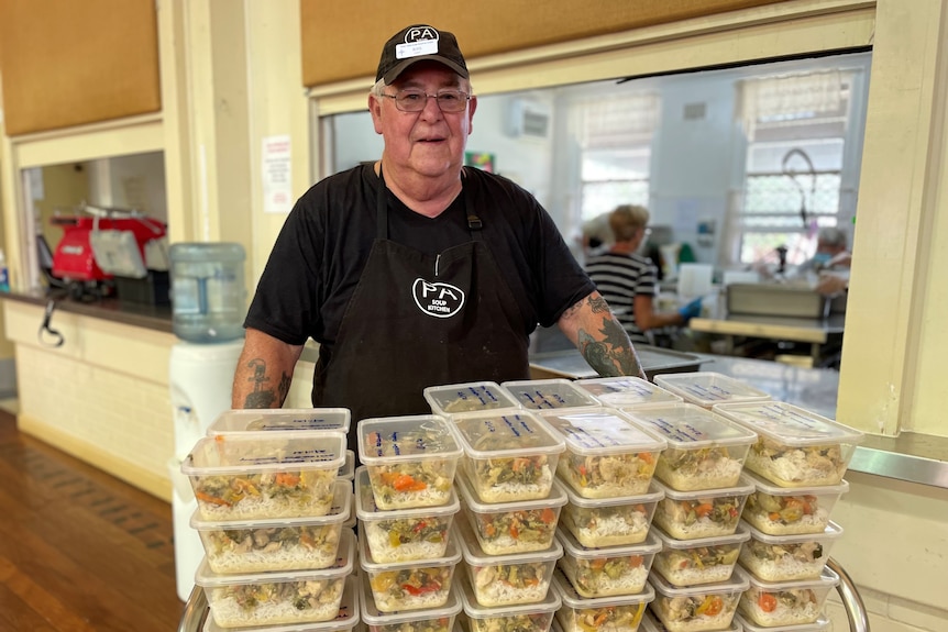An older man wearing a black shirt and cap stands behind a stack of meals in plastic containers.