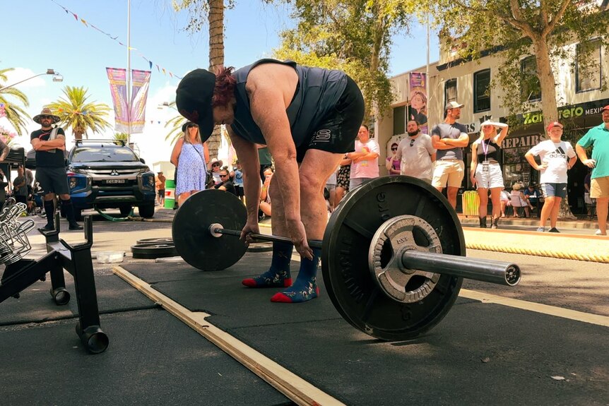 A 73-year-old woman deadlifts with 80kg of weight in the middle of a bitumen street with a crowd.