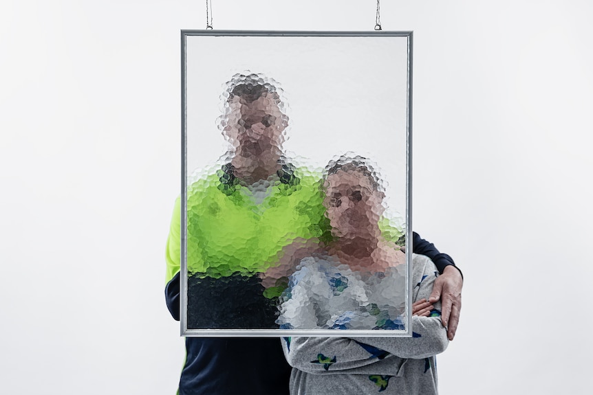A man and woman stand together behind a frame of tempered glass that blurs their faces.