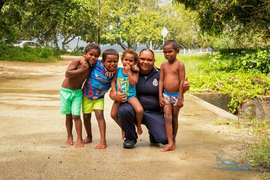 An Aboriginal police liaison officer crouching with four small children all smiling for a photo.