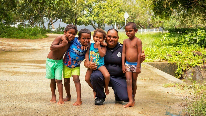 An Aboriginal police liaison officer crouching with four small children all smiling for a photo.