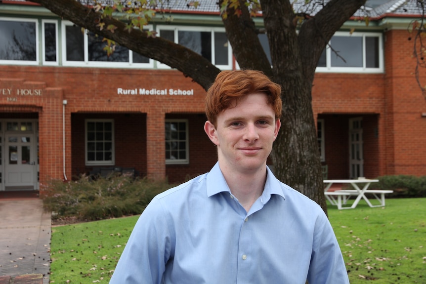 A young man with ginger hair stands outside a brick building bearing the lettering "Rural Medical School".