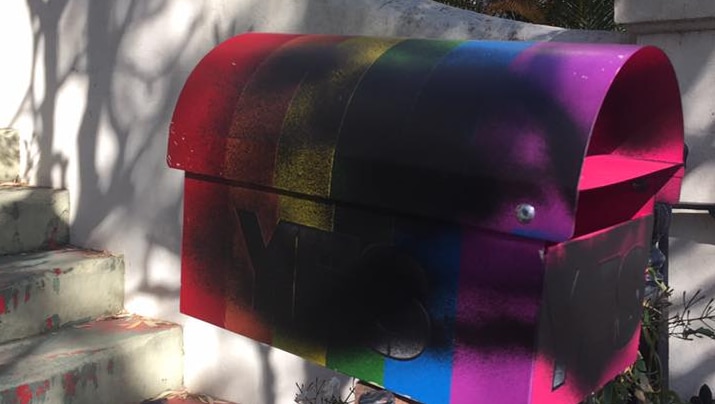 A rainbow-coloured letterbox is defaced with black spray paint.