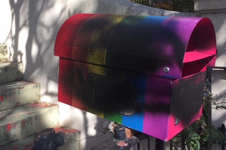 A rainbow-coloured letterbox is defaced with black spray paint.