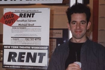 A man with dishevelled brown hair holding a beer stands in front of a sign for Rent that says sold out